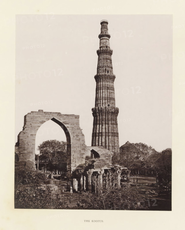 View of the Qutb Minar in Delhi, The Kootur, page, photomechanical print, photographer: Bourne & Shepherd, Delhi, c. 1860 - c. 1877, printmaker, paper, Woodburytype, height 182 mm x width 145 mm, tower, clock tower, monumental adornment, A tall, cylindrical stone tower with carved horizontal bands and decorative balconies rises behind a partially ruined stone archway, both set among trees and scattered stone ruins; the foreground shows smaller stone structures with pillars and vegetation, and a few small human figures near the base of the ruins.