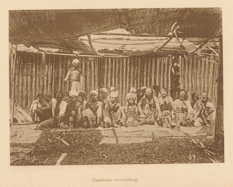 Portrait of unknown persons in costume, some with masks, Gamboeh scene, page, photomechanical print, maker: Christiaan Johan Neeb, c. 1892 - in or before 1897, paper, collotype, photoprint, height 118 mm x width 163 mm, mask, anonymous historical persons depicted in a group, in a group-portrait, stage costume, Indonesia, A sepia-toned photograph showing a group of people seated and kneeling on a floor in front of a wall made of vertical bamboo slats, with a thatched roof overhead supported by wooden poles and beams. The people wear a variety of clothing and head coverings, some holding round handheld fans and some wearing masks or face adornments; one person stands behind the seated group near the center. The ground in front of the group appears covered with woven mats and scattered natural debris, and the scene is framed by the rough wooden structure and roof elements above.