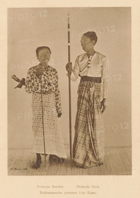 Brahmin priests 1st Caste, Portrait of Pedanda Buddha and Pedanda Siwa, page, photomechanical print, maker: Christiaan Johan Neeb, c. 1892 - in or before 1897, paper, collotype, photoprint, height 166 mm x width 123 mm, historical persons, adult man, Indonesia, A monochrome photograph of two standing people on a plain backdrop, both barefoot on a textured floor. The person on the left is shorter, wearing a patterned long skirt and a floral long-sleeved blouse, holding a thin vertical staff and a small object at their side, with short hair and a neutral facial expression. The person on the right is taller, wearing a light-colored long-sleeved top with decorative fastenings and a checked wrapped lower garment, holding a tall striped pole that reaches above their head, with hair pulled back and looking slightly toward the left. The overall image has a sepia tone and shows both figures in full length.
