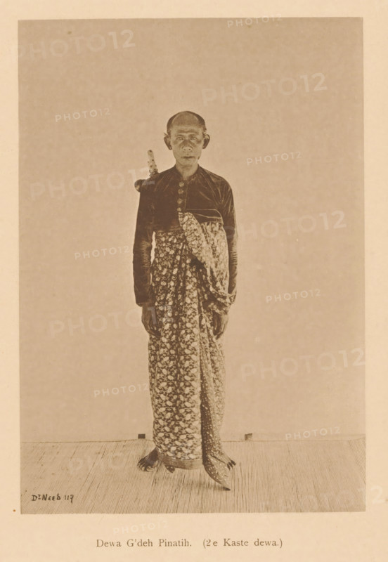 Portrait of Dewa G'deh Pinatih, Dewa G'deh Pinatih (2nd Caste dewa), page, photomechanical print, maker: Christiaan Johan Neeb, c. 1892 - in or before 1897, paper, collotype, photoprint, height 170 mm x width 119 mm, historical persons, adult man, Indonesia, A sepia-toned photograph of a standing person facing forward wearing a patterned wrap skirt and a long-sleeved dark top with decorative buttons, a folded patterned cloth draped across the torso, and a sheathed dagger tucked behind the shoulder; the person has a shaved or closely cropped head, visible ears, and a neutral facial expression, and the image shows a simple plain background and a textured floor beneath their feet.
