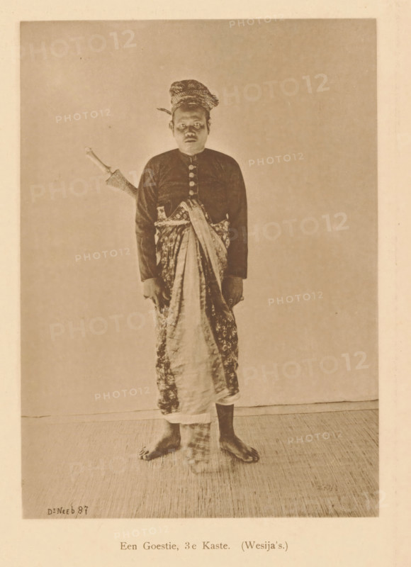 A Goestie, 3rd Caste. (Wesija's), Portrait of an unknown man, page, photomechanical print, maker: Christiaan Johan Neeb, c. 1892 - in or before 1897, Christiaan Johan Neeb, paper, collotype, photoprint, height 170 mm x width 119 mm, historical persons, adult man, Indonesia, A sepia-toned photograph of a barefoot man standing against a plain backdrop, wearing a patterned headwrap, a dark long-sleeved buttoned shirt, and a patterned cloth wrapped around his lower body with one end draped across his torso; a sheathed dagger with a visible handle is tucked at his side, and his hands hang at his sides while his feet rest on a textured floor.