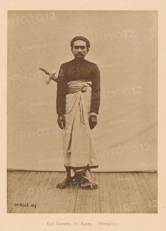 Portrait of an unknown man, A Goestie, 3rd Caste. (Wesija's), page, photomechanical print, maker: Christiaan Johan Neeb, c. 1892 - in or before 1897, Christiaan Johan Neeb, paper, collotype, photoprint, height 170 mm x width 119 mm, historical persons, adult man, Indonesia, A sepia-toned photograph of a person standing against a plain backdrop wearing a dark long-sleeved top and a light wrapped lower garment tied at the waist, with bare feet visible and one foot partially resting on a patterned cloth; the person has short, combed-back hair and a neutral facial expression, and a decorated handle of an object protrudes from the waist area on one side.