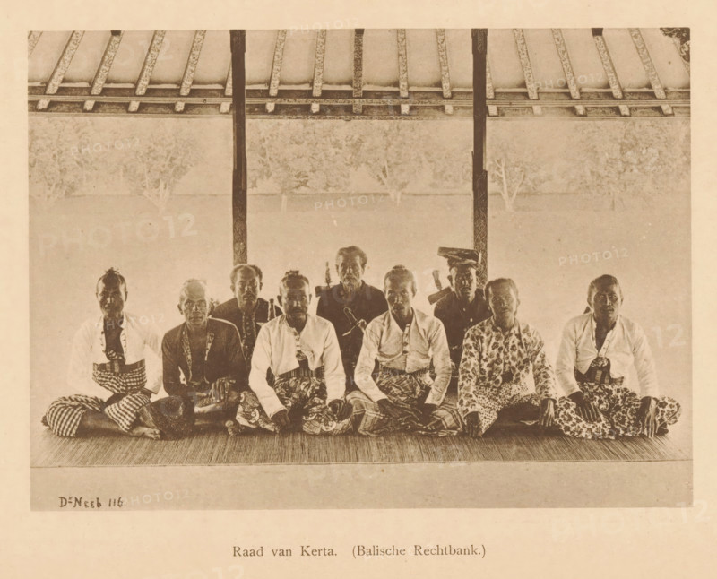 Group portrait of unknown men, the Council of Kerta, Council of Kerta (Balian Court.), page, photomechanical print, maker: Christiaan Johan Neeb, c. 1892 - in or before 1897, paper, collotype, photoprint, height 122 mm x width 167 mm, historical persons, adult man, Indonesia, A sepia-toned photograph showing a row of men seated on a woven mat under a roof with exposed beams and vertical posts, the men wearing patterned cloths wrapped around their lower bodies and a variety of shirts and jackets, some with patterned tops and some plain, several of the men holding or positioned near curved-handled objects, the background behind them appearing light and textured with faint silhouettes of trees, the floor in front of them textured and the scene framed by the roof structure above.