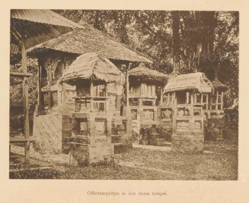 View of sacrificial temples, Sacrificial temples in the Dessa temple, page, photomechanical print, maker: Christiaan Johan Neeb, c. 1892 - in or before 1897, paper, collotype, photoprint, height 124 mm x width 166 mm, trees, places and objects of worship, non-Christian religions, Indonesia, A sepia-toned photograph showing a row of small raised structures with thatched roofs set on carved stone or brick bases, each structure supported by wooden posts and partially enclosed by low ornate balustrades, with larger thatched and corrugated roofs visible behind them, dense foliage of tall trees in the background, and a ground area with grass and scattered stonework in the foreground.