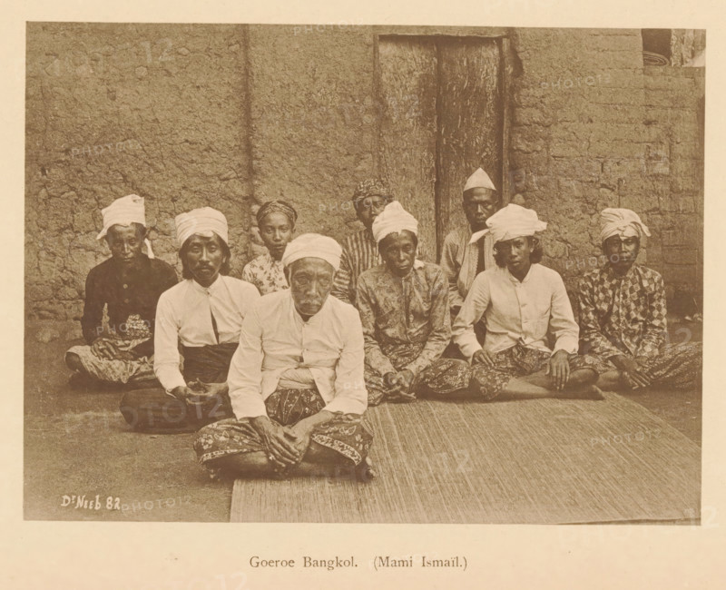 Guru Bangkol. (Mami Ismail), Group portrait of nine unknown men, page, photomechanical print, maker: Christiaan Johan Neeb, c. 1892 - in or before 1897, paper, collotype, photoprint, height 121 mm x width 166 mm, historical persons, adult man, anonymous historical persons depicted in a group, in a group-portrait, Indonesia, A sepia photograph showing a group of people seated on a woven mat in front of a textured wall and a wooden door, most wearing cloth headwraps and patterned lower garments, with several wearing light-colored shirts and some wearing darker patterned tops, all sitting closely together with hands resting in laps or on knees, faces turned toward the camera and showing neutral expressions, the scene framed by the rough wall surface on either side and the mat extending in front of them.