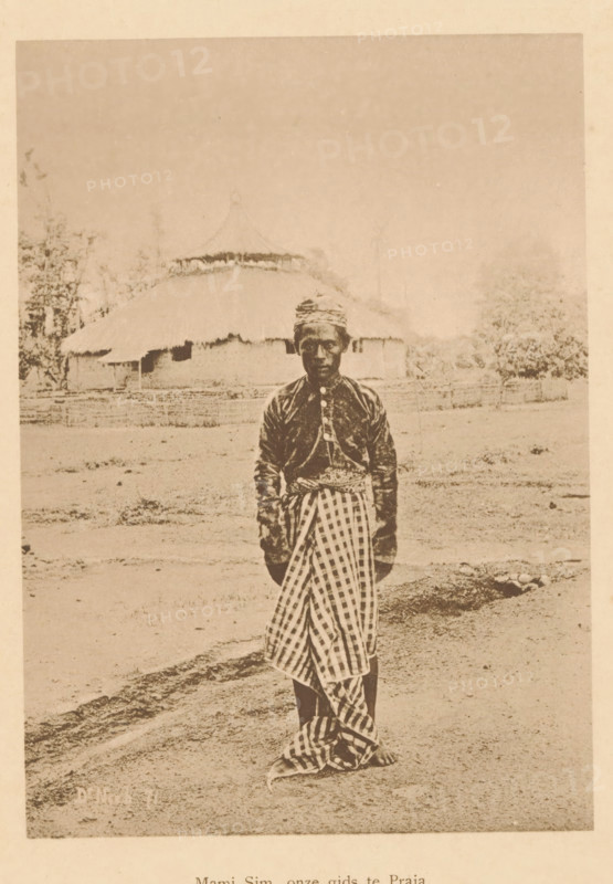 Mami Sim, our guide to Praja, Portrait of Mami Sim, page, photomechanical print, maker: Christiaan Johan Neeb, c. 1892 - in or before 1897, paper, collotype, photoprint, height 169 mm x width 120 mm, historical persons, adult man, Indonesia, A sepia-toned photograph of a person standing outdoors on bare ground, wearing a patterned headwrap, a dark long-sleeved top with buttons down the front, and a checkered cloth wrapped around the lower body, with bare feet visible; behind the person is a low fence and a thatched-roof building with a rounded roofline, surrounded by trees and open space.