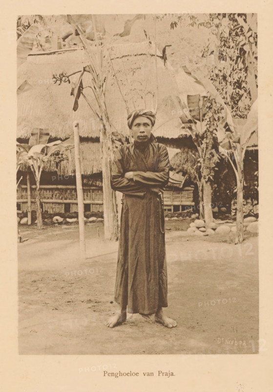 Portrait of an unknown man who is an Islamic teacher, Pengoeloe van Praja, page, photomechanical print, maker: Christiaan Johan Neeb, c. 1892 - in or before 1897, paper, collotype, photoprint, height 161 mm x width 118 mm, historical persons, adult man, Indonesia, A sepia-toned photograph of a barefoot person standing on bare ground with arms crossed, wearing a long dark robe and a wrapped headscarf, positioned centrally in front of a thatched structure and flanked by tall plants with broad leaves; behind them are wooden posts, woven fencing, and clustered stones, with foliage and parts of the thatched roof visible across the upper area of the image.
