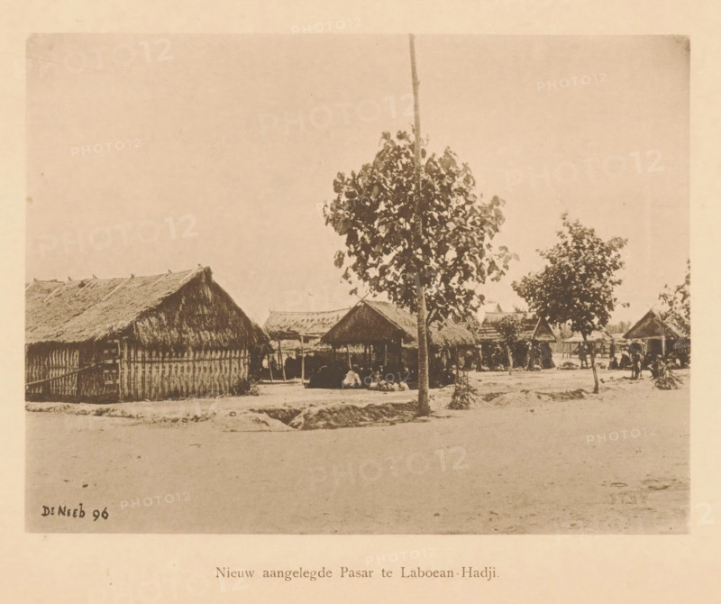Newly constructed Pasar at Laboean-Hadji, View of a market, page, photomechanical print, maker: Christiaan Johan Neeb, c. 1892 - in or before 1897, paper, collotype, photoprint, height 123 mm x width 168 mm, market, Indonesia, A sepia-toned photograph shows a dirt foreground leading to a row of thatched-roof huts and open-sided shelters with woven walls and raised roofs, a tall tree with a slender trunk and a leafy canopy standing near the center, additional smaller trees scattered among the huts, a few people seated or gathered beneath the shelters, simple fencing and posts around some structures, and a mostly clear sky above.