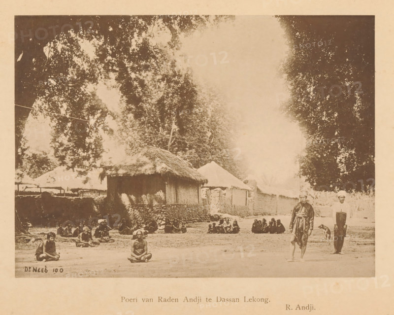 Poeri van Raden Andji te Dassan Lekong, Unknown persons, sitting and standing, at various houses, page, photomechanical print, maker: Christiaan Johan Neeb, c. 1892 - in or before 1897, paper, collotype, photoprint, height 122 mm x width 163 mm, historical persons, other dwelling types, Indonesia, A faded sepia photograph showing a dirt open area in front of simple thatched and walled structures, with large trees rising behind them and a pale sky above; people are seated on the ground in small groups across the foreground and midground, a few individuals sit alone with legs crossed, and two men stand near the right edge beside a dog, one wearing loose garments and the other in lighter clothing; stone or earthen walls and clustered huts form a low row behind the seated figures.
