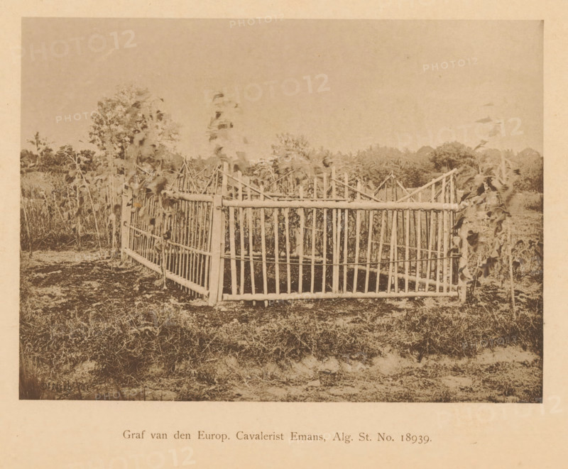 Grave of Emans, Grave of Europ. Cavalryman Emans, General. St. No. 18939, page, photomechanical print, maker: Christiaan Johan Neeb, c. 1892 - in or before 1897, paper, collotype, photoprint, height 122 mm x width 166 mm, landscape with ruins, Indonesia, A sepia-toned photograph shows a fenced enclosure made of vertical and horizontal wooden poles forming a rectangular structure with a gate-like front, set on grassy ground; plants with tall stalks and broad leaves grow around and inside the enclosure, and a tree line is visible in the background beneath a clear sky.