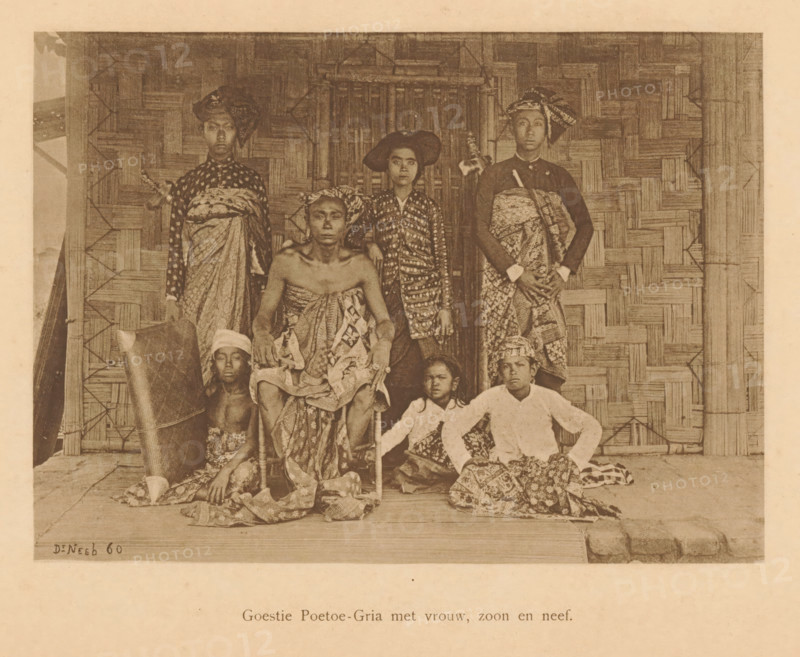 Goestie Poetoe-Gria with wife, son and nephew, Group portrait of a Goestie Poetoe-Gria with six unknown persons, page, photomechanical print, maker: Christiaan Johan Neeb, c. 1892 - in or before 1897, paper, collotype, photoprint, height 166 mm x width 120 mm, historical persons, adult woman, adult man, Indonesia, A sepia photograph shows a group of people arranged in front of a woven bamboo wall and a doorway. Some individuals are seated on a low platform while others stand behind them; several wear patterned cloth wrapped around their bodies and head coverings of different shapes and styles. One seated person in the center sits on a chair with cloth draped over the lap and shoulders, another seated figure beside them wears a lighter-colored top and patterned lower garment, and a few younger figures sit or crouch on the floor with cloths around their waists. The standing figures wear long patterned wraps and head coverings, and one holds a long object across the torso. The floor shows...