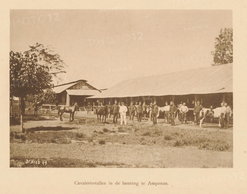 Cavalry stables in the benteng in Ampenan, Group of unknown persons at the cavalry stables in Ampenan, page, photomechanical print, maker: Christiaan Johan Neeb, c. 1892 - in or before 1897, paper, collotype, photoprint, height 120 mm x width 166 mm, historical persons, stall, stable, Lombok, Ampenan, A wide sepia photograph shows a long low building with a pitched roof stretching across the middle of the scene, flanked by taller barnlike structures and trees at the edges; in front of the building a group of men stand in a line, many wearing uniforms and some in lighter clothing, several holding or standing beside horses while others hold bridles or appear to be grooming the animals, and a few figures stand slightly apart near trees and fencing; the foreground is an open grassy area with patches of bare ground.