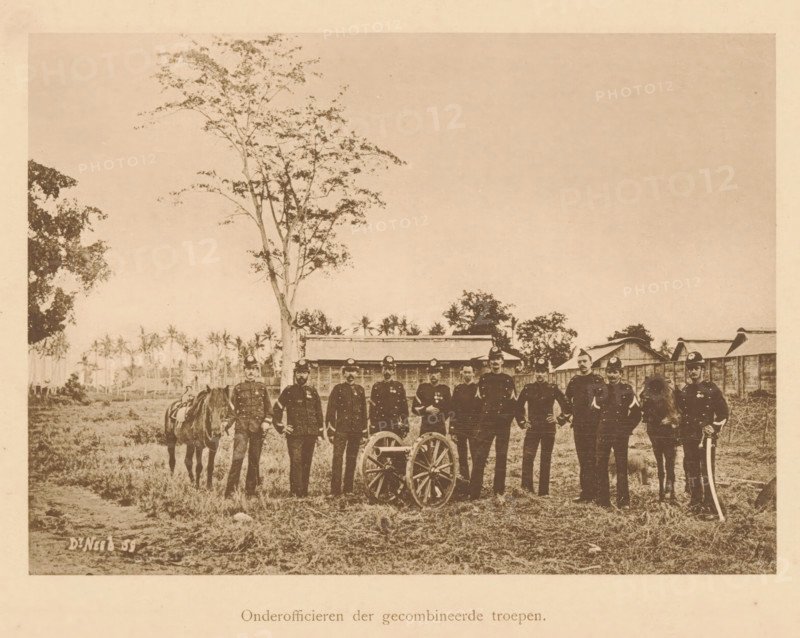 Group portrait of unknown officers near a cannon, Non-commissioned officers of the combined troops, page, photomechanical print, maker: Christiaan Johan Neeb, c. 1892 - in or before 1897, paper, collotype, photoprint, height 122 mm x width 169 mm, firearms: cannon, anonymous historical persons depicted in a group, in a group-portrait, ranks (in the army, navy, air force), A sepia-toned photograph shows a group of uniformed men standing in a line outdoors on grassy ground, some holding objects at their sides and two positioned beside horses, with a small wheeled cart in front of them; behind them are low wooden buildings and scattered trees, including a tall slender tree rising above the group, with a mostly clear sky overhead.