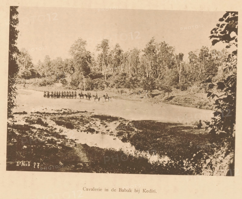 View of the cavalry in a river near Kediri, Cavalry in the Babak near Kediri, page, photomechanical print, maker: Christiaan Johan Neeb, c. 1892 - in or before 1897, paper, collotype, photoprint, height 120 mm x width 167 mm, trees, river, land forces (cavalry, horsemen), Indonesia, A sepia-toned landscape photograph showing a wide shallow river with a line of mounted figures crossing from one bank to the other, leafy trees and dense forest rising behind the far bank, rocky and uneven riverbed in the foreground, dark silhouetted foliage framing the left and right edges of the image, and a small figure seated near the near bank on the right.
