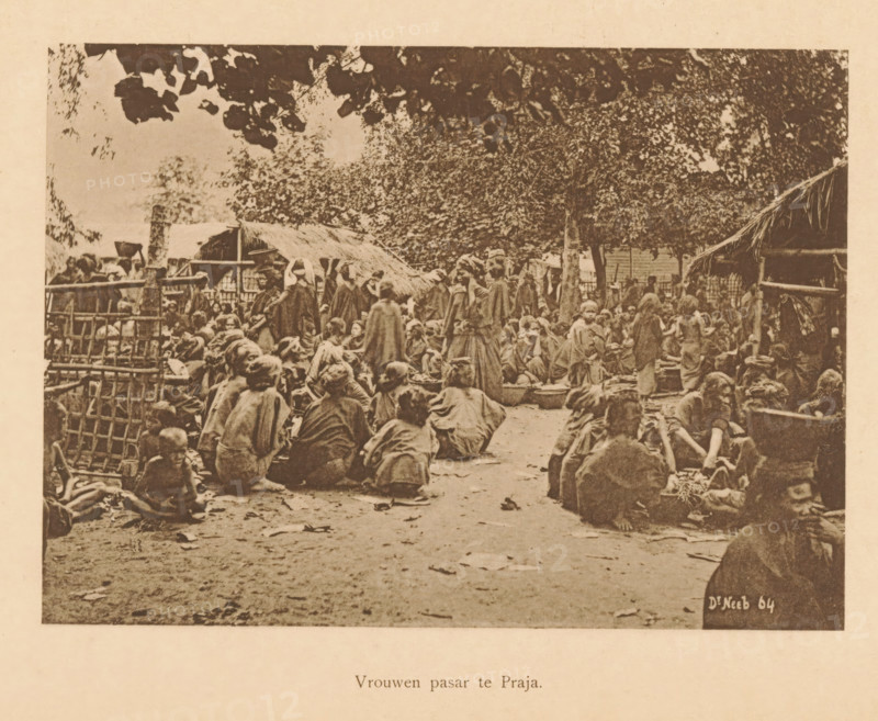 Women's pasar in Praja, Market in Praja, page, photomechanical print, maker: Christiaan Johan Neeb, c. 1892 - in or before 1897, Christiaan Johan Neeb, paper, collotype, photoprint, height 120 mm x width 167 mm, historical persons, market, Indonesia, A sepia-toned photograph showing a crowded outdoor scene with many people seated and standing on a dirt ground, many wearing loose garments and cloaks, some grouped around low bowls and containers, others sitting on the ground or on low stools, with simple thatched shelters and woven fences visible around the perimeter, large leafy trees overhead casting shade, scattered leaves on the ground, and a mix of adults and children occupying the space.