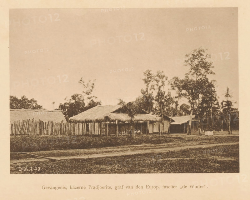 Prison, Pradjoerits barracks, grave of the Europ. huselier 'de Winter', View of the Pradjoerits barracks, page, photomechanical print, maker: Christiaan Johan Neeb, c. 1892 - in or before 1897, paper, collotype, photoprint, height 121 mm x width 168 mm, barracks, Indonesia, A wide sepia-toned photograph showing a row of simple thatched-roof huts and a tall fence made of vertical wooden posts set behind a grassy foreground, with scattered trees of varying heights rising behind and between the structures and a broad expanse of pale sky above, the scene framed by a lighter border around the image.