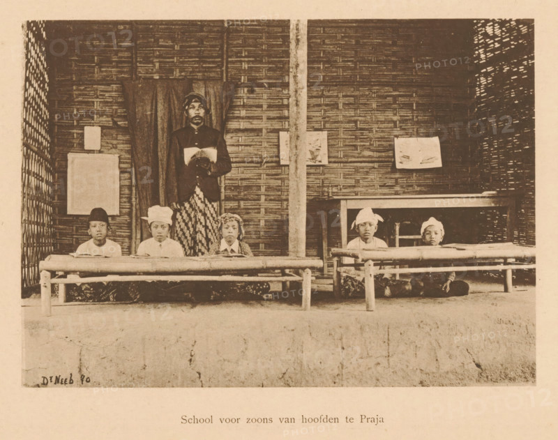 School for sons of headmasters in Praja, View of unknown children and an unknown man in a school, page, photomechanical print, maker: Christiaan Johan Neeb, c. 1892 - in or before 1897, paper, collotype, photogravure, height 118 mm x width 162 mm, historical persons, boy (child between toddler and youth), adult man, Indonesia, A sepia photograph showing a woven bamboo wall with a vertical post near the center and curtains hanging behind a standing man who holds an open book, wearing a dark jacket and patterned lower garment, with five seated boys arranged on low wooden benches in front of him wearing light shirts and various head coverings, some with fabric wraps, and small books or papers on the benches, with a simple wooden railing in front and framed rectangular objects hung on the wall.