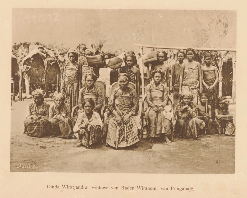 Dinda Wiratjandra, widow of Raden Wiranom, of Pringabaje, Portrait of unknown women and girls with Dinda Wiratjandra, page, photomechanical print, maker: Christiaan Johan Neeb, c. 1892 - in or before 1897, paper, collotype, photogravure, height 120 mm x width 165 mm, Person, historical persons, adult woman, adolescent, young woman, maiden, girl (child between toddler and youth), Indonesia, A sepia-toned photograph showing a group of people arranged in rows outdoors in front of tall leafy plants and a simple wooden structure, with several seated individuals wearing patterned cloth wrapped around their bodies and headscarves, and others standing behind them carrying woven baskets and bundles on their heads or shoulders; the seated row includes older women with folded hands and a few younger individuals seated on the ground, while the standing row shows women in wrapped garments and a couple of children, all facing the camera with neutral expressions and the ground in front of them visibl...