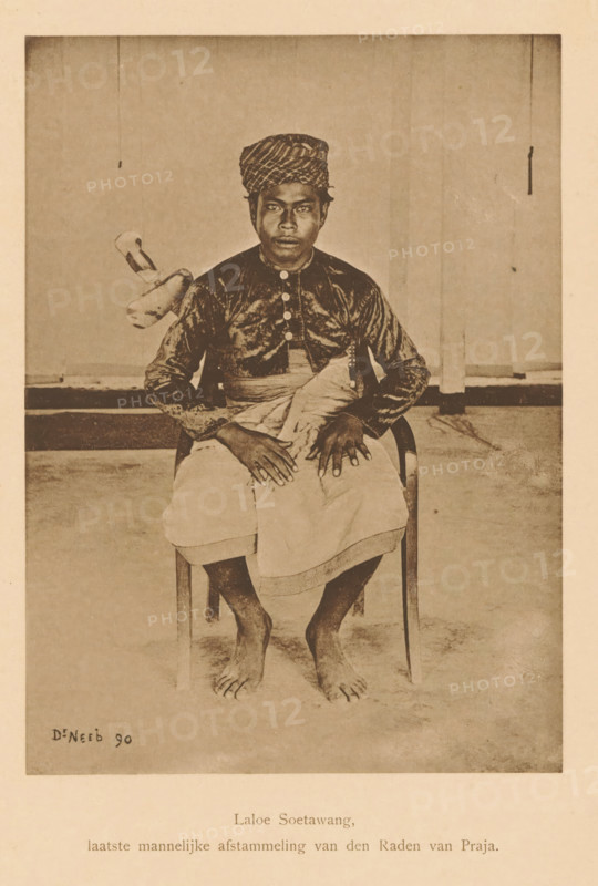 Laloe Soetawang, last male descendant of the Raden van Praja, Portrait of Laloe Soetawang, page, photomechanical print, maker: Christiaan Johan Neeb, c. 1892 - in or before 1897, paper, collotype, photoprint, height 170 mm x width 123 mm, Person, historical persons, adult man, Indonesia, A sepia-toned photograph of a barefoot man seated on a wooden chair facing forward, wearing a patterned headwrap and a dark, long-sleeved top with a row of buttons down the front, a light cloth wrapped around his waist, hands resting on his lap with fingers spread, bare feet visible on the floor, a plain backdrop behind him with a hanging cord and a small object suspended to one side, and a small signature visible in the lower corner of the image.