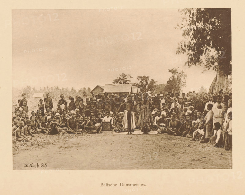 Balian Dancing Girls, Group of unknown persons watching two dancers, page, photomechanical print, maker: Christiaan Johan Neeb, c. 1892 - in or before 1897, paper, collotype, photoprint, height 120 mm x width 165 mm, adult man, dancing, anonymous historical persons depicted in a group, in a group-portrait, Indonesia, A wide sepia photograph shows a large crowd of people gathered outdoors with trees and simple thatched-roof huts in the background, many seated on the ground and others standing around a central open space where two women wearing patterned skirts and decorative tops stand facing forward, a few seated figures near them appear to hold drums or boxes, the crowd includes men, women, and children in traditional and simple clothing, and a large tree with hanging leaves is visible on the right edge of the scene.