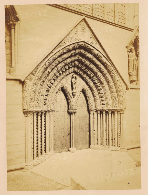 Portal of Lichfield Cathedral, photograph, page, photographer: John Benjamin Stone, Lichfield, c. 1865 - in or before 1870, Staffordshire, paper, albumen print, height 177 mm x width 128 mm, church (exterior), parts of church exterior and annexes: portal, parts of church exterior and annexes: tympanum, Lichfield Cathedral, A sepia-toned photograph of a carved stone doorway featuring a pointed arch with multiple concentric archivolts decorated with carved foliage and small sculpted heads, a central sculpted figure standing above double wooden doors studded with decorative rivets, slender clustered columns on either side with decorated capitals, flat stone walls surrounding the entrance, and a sculpted standing figure in a recessed niche to the right of the doorway.