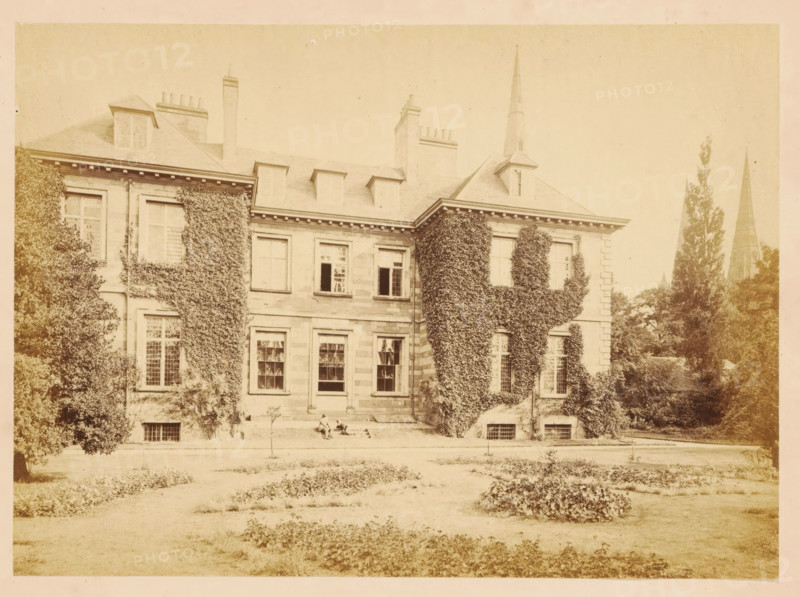 View of Bishop's Palace at Lichfield, photograph, page, photographer: John Benjamin Stone, Lichfield, c. 1865 - in or before 1870, John Benjamin Stone, photographer, Staffordshire, paper, albumen print, height 126 mm x width 173 mm, facade (of house or building), A wide pale photograph of a large stone building with a pitched roof and several chimneys, its facade partly covered in climbing ivy, with multiple tall windows on two levels and dormer windows in the roof; a trimmed lawn and garden beds lie in the foreground, and there are tall trees near the building and two narrow pointed church spires rising behind the trees to the right.