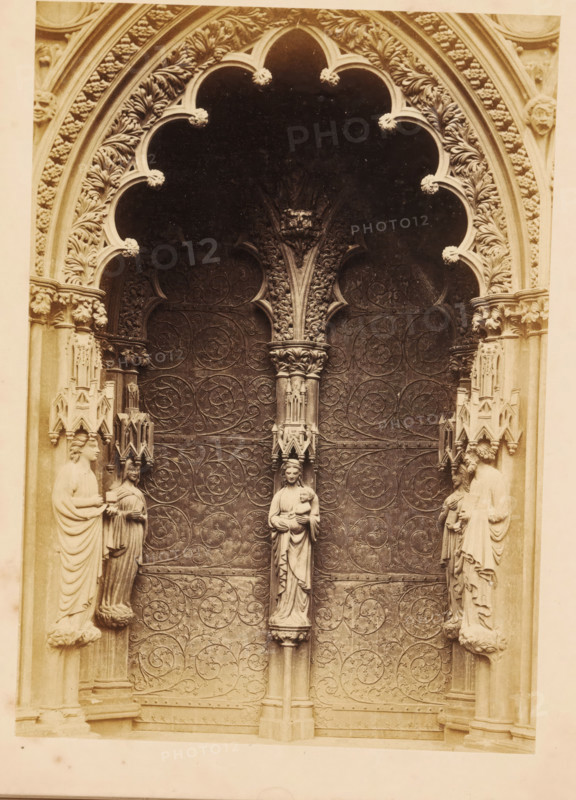 Portal of Lichfield Cathedral, photograph, page, photographer: John Benjamin Stone, Lichfield, c. 1865 - in or before 1870, Staffordshire, paper, albumen print, height 180 mm x width 127 mm, church (exterior), parts of church exterior and annexes: portal, An ornate stone doorway with a scalloped arch richly carved with foliage motifs frames a dark recessed area; the door surface is covered in swirling vine-like relief patterns. Flanking the doorway are carved columnar figures standing on small pedestals, each draped in flowing robes and positioned within decorative niches. A central slender column rises between the door panels, topped with an elaborately carved capital and a statue of a robed female figure holding a small child. Additional small sculptural canopies and intricate architectural details adorn the jambs and capitals on either side. The image has a monochrome, sepia-toned appearance.