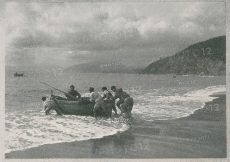 Seascape with fishermen, Italian fischer, page, photomechanical print, monogram: 'BX', photographer: W. Schmidt-Diehler, cliché maker: unknown, c. 1898 - in or before 1903, paper, autotypie, height 130 mm x width 125 mm, fisherman, sea (seascape), A grayscale scene of a shoreline with a group of people pushing a small boat from shallow surf toward the beach, their clothing appearing dark against the lighter foam of the waves; the boat sits at the waterline with a long pole extending from it, and another small vessel is visible farther out on the water; the sky above is filled with heavy, textured clouds, and a dark, sloping landmass frames the right side of the horizon while gentle waves wash onto the sand in the foreground.