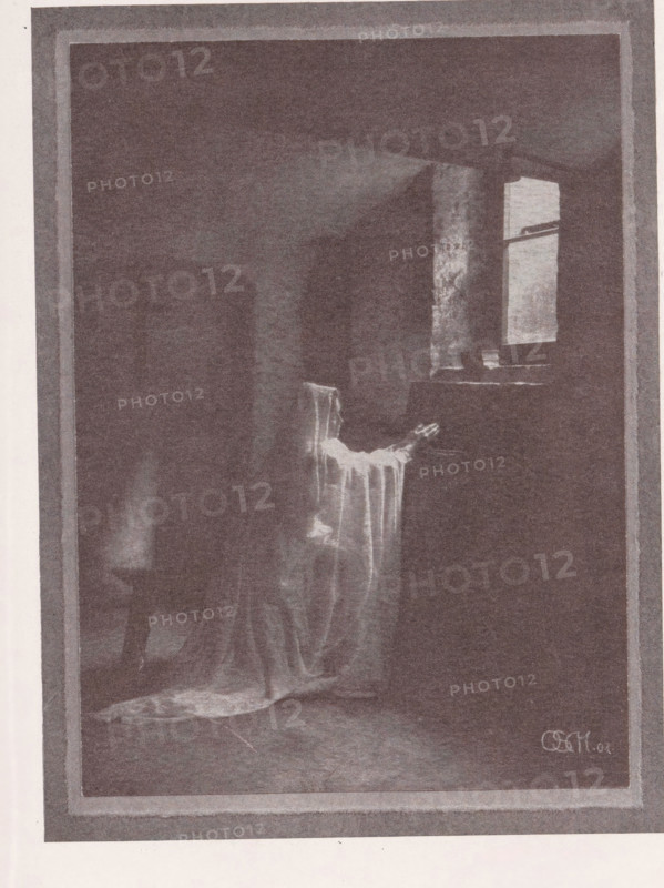 Portrait of an unknown woman depicted as a penitent, Büsserin, page, photomechanical print, inscription: 'Photographische Mitteilungen XL', photographer: Otto Scharf, Krefeld (possibly), cliché maker: anonymous, c. 1898 - in or before 1903, North Rhine-Westphalia, paper, autotypie, height 174 mm x width 129 mm, historical persons - BB - woman, A dim interior scene framed by a wide border shows a draped chair near a window, with light falling through the window onto a portion of the chair and floor; the chair is covered with a flowing, translucent fabric that cascades to the floor, creating soft folds and a small pooled area; a narrow table or ledge sits beneath the window with a few indistinct objects on it; the room beyond the chair is dark and shadowed, with a suggestion of other furniture shapes obscured by low light; the overall image has a grainy, monochromatic appearance.