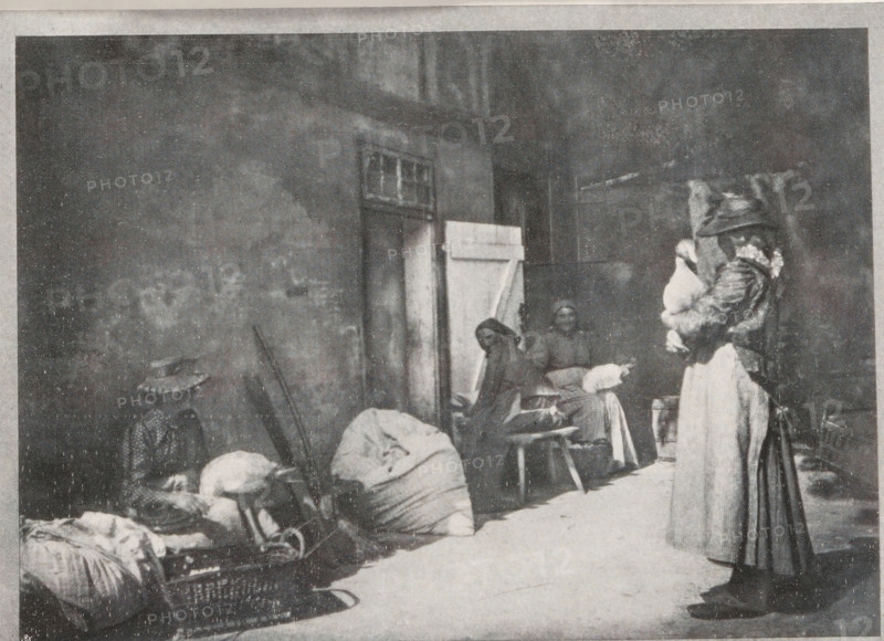 Gänse rupfen, Plucking geese, page, photomechanical print, inscription: 'Berlin', monogram: 'BX', inscription: 'Photographische Mitteilungen XL', photographer: Hildegard Lehnert, cliché maker: unknown, c. 1898 - in or before 1903, paper, autotypie, height 128 mm x width 176 mm, geese (domestic waterbirds), A grainy black and white street scene shows a tall wall with a doorway partly covered by a half-open door and a small window above it, against which several people and piles of large wrapped bundles are arranged; on the left a seated figure wears a wide-brimmed hat and sits beside sacks and a basket, near a person bent over the bundles, while toward the center two seated women are positioned on simple stools with one facing forward and the other turned slightly, and on the right a standing woman wearing a long skirt and a coat with a hood or head covering holds a bundled child against her chest.