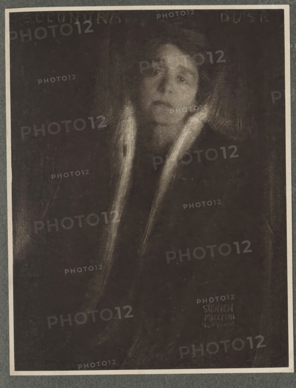 Eleonore Duse, Portrait of Eleonore Duse, page, photomechanical print, inscription: 'Photographische Mitteilungen XL', photographer: Edward Steichen, New York (city) (possibly), cliché maker: anonymous, c. 1898 - in or before 1903, North America, paper, photoaquatint, height 181 mm x width 140 mm, Person, Eleonora Duse, historical persons - BB - woman, A dark, grainy portrait of a person with a pale face and an expressionless gaze, framed by soft, light strokes suggesting hair and a collar or scarf that extends downwards, set against a deep, nearly black background with faint vertical and diagonal smudges.