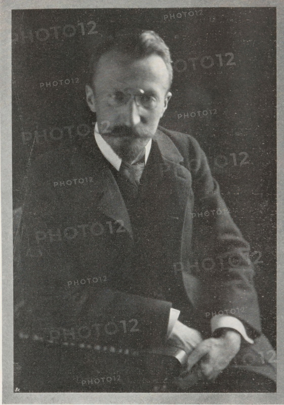 Field camera, page, photomechanical print, c. 1898 - in or before 1903, paper, autotype, height 76 mm x width 133 mm, camera, photography, A black-and-white portrait of a seated man wearing a dark suit with a vest and a light-colored shirt collar, showing a mustache and beard, round glasses, neatly combed hair, and his hands resting in his lap while holding a rolled object.