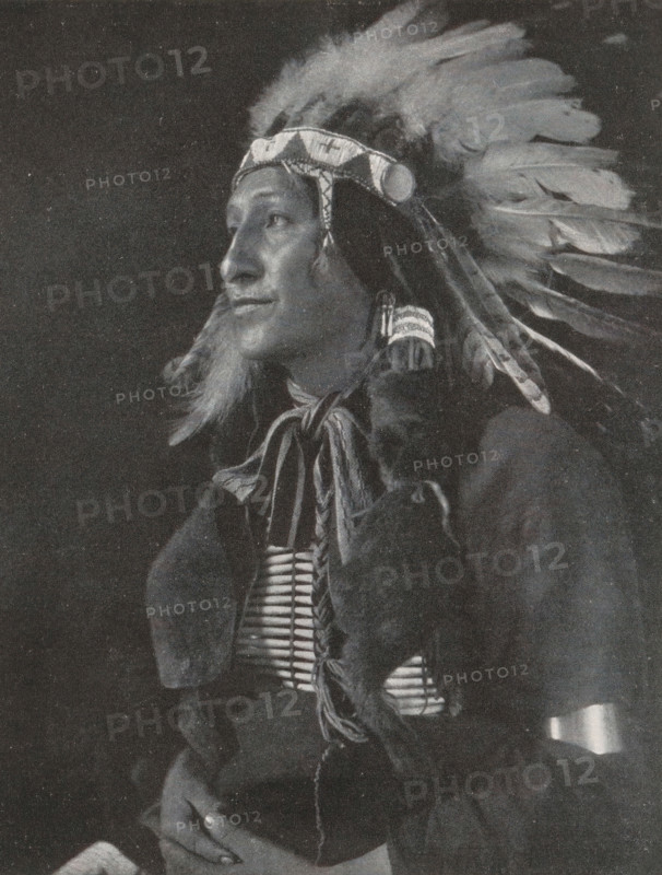 Portrait of an unknown man, The Black Fox, page, photomechanical print, monogram: 'MR&C', photographer: Gertrude Käsebier, New York (city), cliché maker: Meisenbach, Riffarth & Cie., c. 1898 - in or before 1903, North America, paper, autotypie, height 166 mm x width 129 mm, historical persons, A black and white portrait of a person wearing a feathered headdress with a decorative headband and long feathers extending backward, the person shown in three-quarter profile looking to the side, with long hair and braided elements, a beaded or bone chest ornament visible beneath layered clothing, fur or textured fabric draped over the shoulders, hands resting together in the lower frame, and a dark background surrounding the figure.