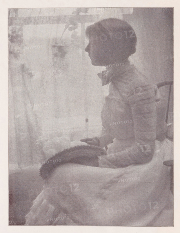 Julia, Portrait of an unknown woman, page, photomechanical print, monogram: 'MR&C', photographer: Gertrude Käsebier, New York (city), cliché maker: Meisenbach, Riffarth & Cie., c. 1898 - in or before 1903, North America, paper, autotypie, height 172 mm x width 130 mm, historical persons - BB - woman, A grainy monochrome photograph showing a woman in profile seated on a cushioned surface facing left, wearing a high-collared blouse with a bow at the neck and a skirt, her hair styled short and rounded; she holds a round hat in her lap and a delicate object with a thin handle is visible near her hand; behind her a light window with semi-sheer curtains and vague floral shapes is visible, and a simple wooden chair back appears beside her.