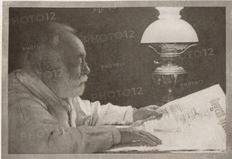 Bei der lampe, Portrait of an unknown man, page, photomechanical print, monogram: 'BX', inscription: 'Copenhagen', inscription: 'Photographische Mitteilungen XL', photographer: Niels Adam Fischer, cliché maker: unknown, c. 1898 - in or before 1903, paper, autotypie, height 125 mm x width 181 mm, historical persons, A grayscale image of an elderly man in profile leaning forward with a beard and glasses, wearing a loose garment and resting his forearms on a surface while holding a large folded paper, and a decorative lamp with an ornate base and a rounded glass shade positioned beside him.