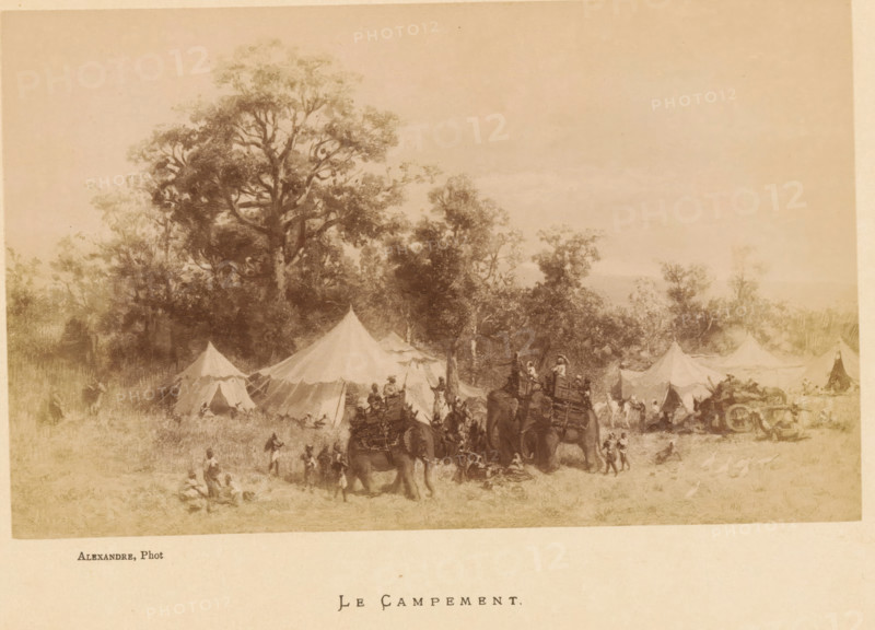Le Campement, Photo reproduction of Le Campement by Jean Baptiste Robie, photograph, reproduction, page, photographer: Alexandre, after painting by Jean Baptiste Robie, c. 1878 - in or before 1883, paper, albumen print, height 113 mm x width 182 mm, behind cover., tent-camp, hunt, predatory animals (fox, lion), A wide sepia-toned scene shows a campsite set at the edge of a wooded area with tall trees rising behind a row of canvas tents of varying shapes and sizes; in front of the tents people move about while figures ride and lead pack animals loaded with gear, and other people stand or sit on the grassy ground, with clusters of activity near the animals and tent openings.