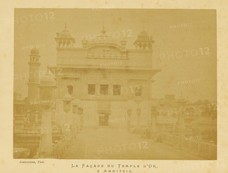La Façade du Temple d'Or, a Amritsir, View of the Harmandir Sahib in Amritsar, photograph, page, photographer: Alexandre, Amritsar, c. 1880 - in or before 1885, Alexandre, photographer, paper, albumen print, height 132 mm x width 177 mm, behind cover., temple, shrine, non-Christian religions, Harmandir Sahib, A faded sepia photograph of a large ornate building with domed roofs and decorative turrets set behind a foreground walkway with low balustrades, showing arched windows and recessed doorways on the building facade, a tall slender tower with a small dome visible to the left, layered rooflines with projecting eaves and sculpted parapets, stone finials and small pavilion-like structures on the roof, and a broad open courtyard or approach leading up to the main entrance.