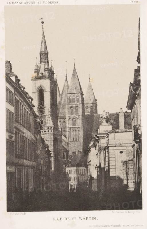 Rue de St. Martin, View of the Rue Saint-Martin in Tournai, Belgium, page, photomechanical print, inscription: 'PHOTOLITHOGRAPHIE PROCÉDÉS ASSER ET TOOVEY', number: 'Pl. XIX', photographer: L. Duchatel, Tournai (Hainaut), cliché maker: Simonau & Toovey (possibly), Brussels, c. 1859 - in or before 1864, clichémaker, printer (person), paper, photolithography, height 216 mm x width 142 mm, street, church (exterior), Cathedral of Our Lady (Tournai), A narrow street flanked by tall buildings leads toward a large stone cathedral with multiple pointed towers and spires rising above the rooftops; the cathedral facade shows arched windows, a large clock face set in a tower, and decorative stonework, while the street-level buildings display rows of windows, balconies, and cornices, with a lamppost visible on one side and shadowed figures and darker foreground tones along the roadway.
