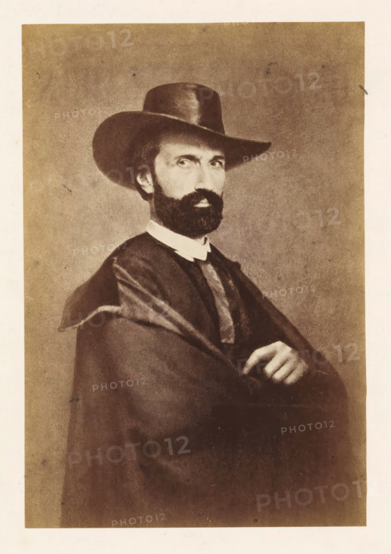Portrait of Antoine-Joseph Wiertz, photograph, page, photographer: attributed to Edmond Fierlants, c. 1855 - in or before 1865, Attributed to, paper, albumen print, height 169 mm x width 114 mm, Photo opposite title page., Person, Antoine Joseph Wiertz, historical persons, A sepia-toned portrait of a man shown from the waist up wearing a wide-brimmed hat and a dark cloak draped over his shoulders, with a white collar visible at his neck; he has a full dark beard and mustache, short hair peeking beneath the hat, and a serious expression while his arms are crossed with one hand resting on the other.