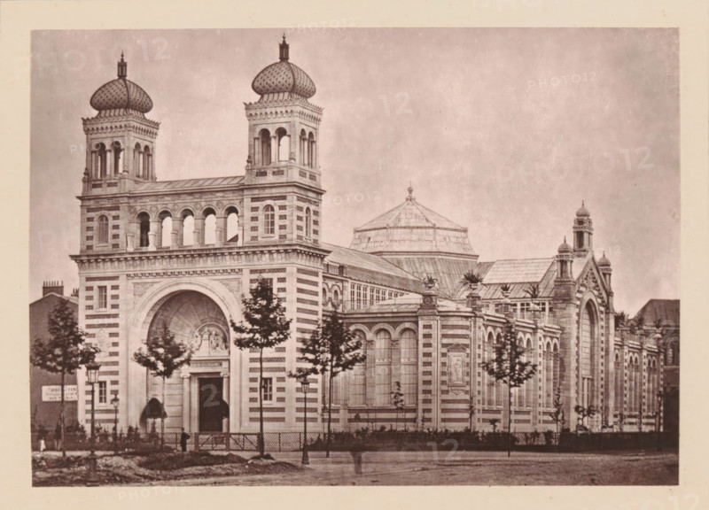 Lille Nouveau, Le Palais Rameau, View of the Palais Rameau in Lille, page, photomechanical print, inscription: 'Ou aura Lieu, le 1er avril, l'exposition des artsindustriels', inscription: 'Le Nord Contemporain, Revue Artistique & Archéologique', c. 1878 - in or before 1882, paper, Woodburytype, height 135 mm x width 190 mm, Print on third unnumbered page after page 96., palace, Lille, A sepia-toned photograph of a large ornate building with striped stonework and arched windows, featuring two square towers topped with bulbous domes and open archways, a central rounded arched entrance with carved relief above, and a low dome visible behind the main roof. The facade includes decorative cornices, pilasters, and sculpted panels, with rows of tall arched windows and smaller rectangular windows. Trees line the front of the building, partially obscuring the lower facade, and lamp posts and a fence are visible along the sidewalk. The image has a wide uniform sky above and a pale border around t...