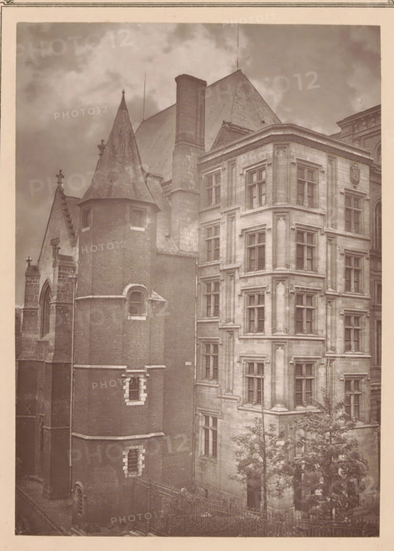Lille, l'hotel-de-ville, Palais Rihour in Lille, page, photomechanical print, inscription: 'Journal le Nord Contemporain, No 13, année 1882', inscription: 'Le Nord Contemporain, Revue Artistique & Archéologique', inscription: 'La partie du onument appelée 'Le Conclave'', c. 1878 - in or before 1882, paper, Woodburytype, height 124 mm x width 152 mm, Print on fifth unnumbered page after page 208., palace, A sepia-toned photograph showing two adjoining historic buildings with varied architectural features. On the left a cylindrical brick turret with narrow arched windows and stone trim rises to a conical roof topped by a finial, adjoining a steeply pitched roof and a tall chimney stack. On the right a stone façade with multiple recessed window bays, decorative moldings, and corner pilasters presents stacked rectangular windows and ornate stonework around an entrance partially obscured by trees. A wrought-iron fence runs along the foreground, and cloudy sky fills the upper background.