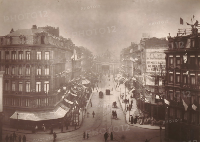 Lille, vue de la Rue de la Gare, View of the Rue Faidherbe with the train station in Lille, page, photomechanical print, inscription: 'Le Nord Contemporain, Revue Artistique & Archéologique', photographer: Lemercier, cliché maker: Mallart, c. 1878 - in or before 1882, paper, Woodburytype, height 151 mm x width 212 mm, Print on third unnumbered page after page 240., city-view in general, 'veduta', street, A wide street scene viewed from above shows rows of multi-story buildings with mansard roofs and dormer windows lining both sides, shop awnings and flags projecting over the sidewalks, scaffolding and rooftop structures on some buildings, and a crowd of people on the sidewalks and walking in the street. Horse-drawn carriages and a few early automobiles move along tracks embedded in the pavement, while lampposts stand along the roadway. The far end of the street is framed by a large archway or gateway between buildings, and the sky above is overcast.