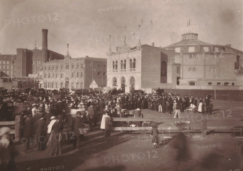 Roubaix, l'Hippodrome a la sortie des sociétés chorales, View of the Grand Place in Roubaix with visitors and a church choir, page, photomechanical print, inscription: 'Pendant les fêtes des 13, 14 et 15 Août', inscription: 'Le Nord Contemporain, Revue Pittoresque', photographer: Lemercier, cliché maker: Ferrand, c. 1878 - in or before 1882, Lemercier, Ferrand, photographer, cliché maker, paper, Woodburytype, height 151 mm x width 214 mm, Print on third unnumbered page after page 272., square, circus, choir, A wide sepia photograph shows a crowded outdoor scene in front of large brick buildings with varied architectural details; a grand façade with arched windows and decorative pinnacles stands near a round building with a domed roof and a flag on top, while adjacent structures include a tall tower and multi-story brick blocks. The foreground is filled with many people wearing period clothing, including hats, coats, long dresses, and aprons, some pushing or standing near small wheeled ...