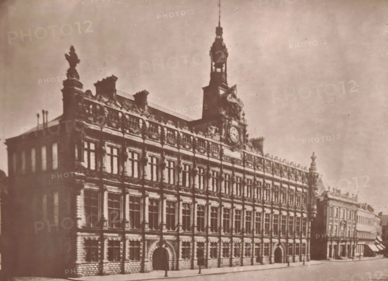 View of the Valenciennes town hall, L'Hotel-de-Ville de Valenciennes, page, photomechanical print, inscription: 'Le Nord Contemporain, Revue Artistique & Archéologique', photographer: Lemercier, Valenciennes, cliché maker: Lemercier, c. 1878 - in or before 1882, Lemercier, photographer, cliché maker, paper, Woodburytype, Woodburytypepie, height 142 mm x width 194 mm, Print on third unnumbered page after page 48., townhall, A sepia-toned photograph of a large ornate building seen from an angle showing its front and side façades, with multiple rows of tall rectangular windows set in decorative stonework and arched doorways at street level, a detailed cornice and sculptural roofline with statues and ornamental elements, and a central tower rising above the roof topped by a spire and housing a circular clock face; the street in front is mostly empty with a few indistinct figures near the building and adjacent lower buildings extending along the right side of the image.