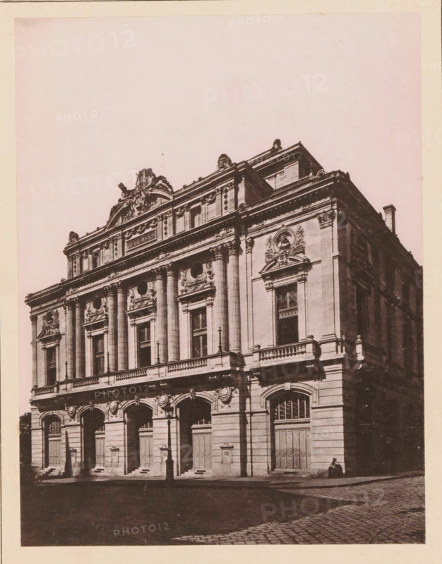 Rouen, Le Nouveau Théatre des Arts, View of the Théatre des Arts in Rouen, page, photomechanical print, inscription: 'Le Nord Contemporain, Revue Historique & Archéologique', inscription: 'Journal le Nord Contemporain, No 20, année 1882.', photographer: Bruckmann, cliché maker: Ferrand, c. 1877 - in or before 1882-10-31, paper, collotype, photoprint, height 209 mm x width 159 mm, Print on fourth unnumbered page after page 320., theater (building), A large ornate stone building fills the frame, showing a decorated facade with tall columns, arched doorways at ground level, and sculpted reliefs and medallions above the windows; a balustraded balcony runs along the middle level, and elaborate cornices and attic sculptures crown the roofline; a solitary streetlamp stands in front of the entrance area, and a person sits on the pavement near the building corner; the foreground shows a cobbled street and a shadowed sidewalk.