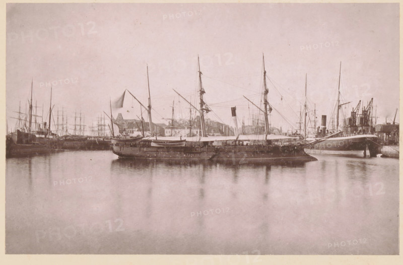 La Lionne, Canonnière de l'État, View of La Lionne near Calais, a gunboat of France, page, photomechanical print, inscription: '(en rade de Calais)', inscription: 'Le Nord Contemporain, Revue Pittoresque', photographer: NC, Calais, cliché maker: Luzzato, Dunkirk, c. 1877 - in or before 1882-12-15, NC, Luzzato, photographer, cliché maker, paper, collotype, photoprint, height 160 mm x width 251 mm, Print on third unnumbered page after page 368., sailing-ship, sailing-boat, firearms: cannon, A wide harbor scene showing several sailing vessels and steamships moored along a quay, with tall masts, rigging, and spars rising against a pale sky, a larger ship in the foreground with lifeboats and canvas covers on its deck, reflections of the ships visible on calm water, a line of warehouses and cranes along the shore behind the vessels, and an overall muted, monochrome tone.