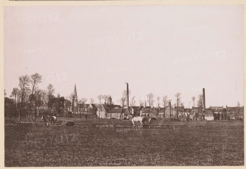 Un Paysage du Nord, Two farmers with horses working in the fields in Northern France, page, photomechanical print, inscription: 'Le Nord Contemporain, Revue Artistique & Pittoresque', photographer: NC, Northern France, cliché maker: C. Becquereau, Saint-Omer, c. 1877 - in or before 1882-12-31, paper, collotype, photoprint, height 159 mm x width 237 mm, Print on fifth unnumbered page after page 384., farmers, landscapes, horse, soil cultivation, A wide, sepia-toned landscape showing a large open field in the foreground with short grass, a few horses and people near the center and left edges, and a line of tall, leafless trees along the middle ground. Behind the trees a collection of buildings is visible, including a church with a pointed spire, other pitched-roof structures, and two tall vertical chimneys. The sky above is pale and mostly empty, occupying the upper portion of the image.
