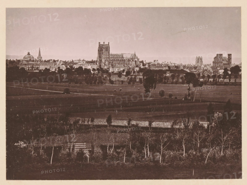 Vue Générale de Saint-Omer, View of Saint-Omer, page, photomechanical print, inscription: 'Le Nord Contemporain, Revue Historique & Pittoresque', photographer: NC, Saint-Omer, cliché maker: C. Becquereau, c. 1878 - in or before 1883-02-05, paper, collotype, photoprint, height 175 mm x width 238 mm, Print on fifth unnumbered page after page 32., prospect of city, town panorama, silhouette of city, A wide sepia-toned cityscape seen from a distance with a foreground of darker fields and rows of leafless trees, a midground of lighter cultivated land with scattered trees and a few small structures, and a background skyline dominated by large historic buildings including a cathedral with tall towers and intricate stonework, a domed building, a slender spire, and several multi-story stone structures, all beneath a pale sky.