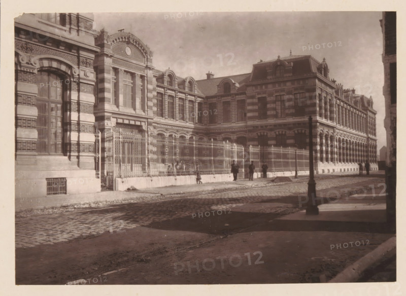 Lille, La Nouvelle Faculté de Médicine, View of Place de la République in Lille during a market, page, photomechanical print, inscription: 'Le Nord Contemporain, Revue Artistique & Archéologique', inscription: 'Elevée par MC Batteur, Architecte', inscription: 'Journal le Nord Contemporain, No 18, année 1882.', photographer: Lemercier, cliché maker: Ferrand, c. 1877 - in or before 1882-09-30, paper, Woodburytype, height 152 mm x width 212 mm, Print on fifth unnumbered page after page 288., market, university (building, education, tuition), A sepia-toned photograph of a large ornate brick and stone building with tall arched windows, decorative cornices, and a clock set in an elaborately carved pediment near the roofline, a metal fence with ornate vertical bars runs along the sidewalk in front of the building, several people stand or walk near the fence and along the cobblestone street, a lamppost stands near the curb casting a shadow on the pavement, the foreground shows a rough dirt and...