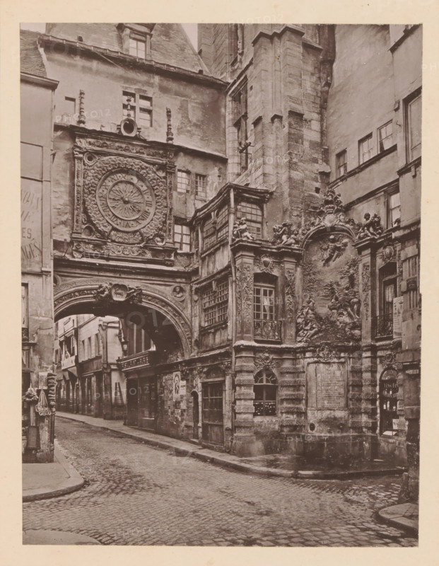 Rouen, La Grosse Horloge, View of the Gros-Horloge in Rouen, page, photomechanical print, inscription: 'Le Nord Contemporain, Revue Artistique & Archéologique', inscription: 'Journal le Nord Contemporain, No 27, année 1883.', photographer: NC, c. 1878 - in or before 1883-02-20, paper, collotype, photoprint, height 221 mm x width 168 mm, Print on third unnumbered page after page 48., tower, clock tower, monumental adornment, Gros-Horloge, A narrow cobblestone street lined with tall, weathered buildings leads under a large ornate stone archway attached to a facade featuring an elaborate circular clock surrounded by carved floral and scrollwork motifs. Above and beside the arch are multi-paned windows and sculpted decorative elements including relief panels with figures and cherubs, carved pilasters, and balustraded window boxes. The stone surfaces show aged textures and darkened areas, and a small inset doorway and arched window occupy the ground level beneath the relief. A portion of th...